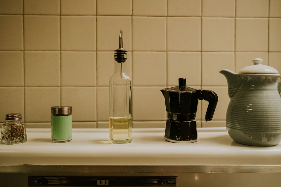 A collection of vintage kitchen essentials on a tiled countertop, featuring a coffee pot, oil dispenser, and salt shaker.